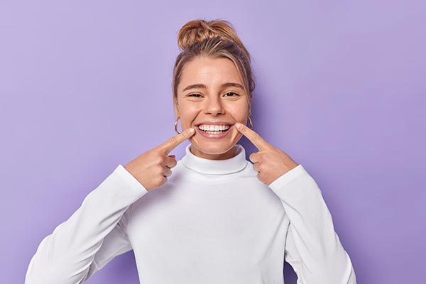Woman with straight teeth holding her face with both hands, smiling broadly against a purple background.