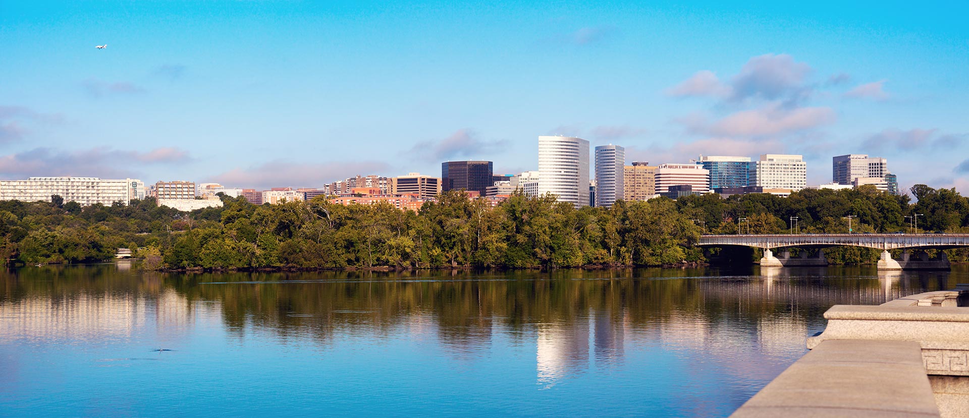The image shows a serene cityscape with a body of water reflecting the skyline, a bridge spanning across the water, and buildings visible in the background under a clear blue sky.