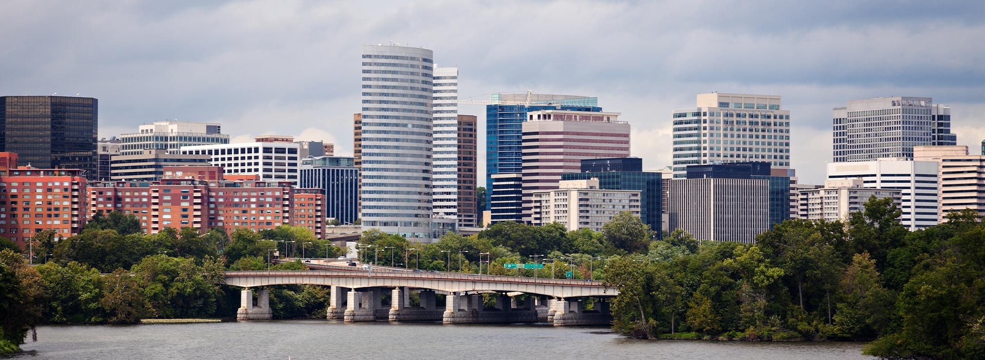 The image shows a city skyline with tall buildings, including a prominent bridge, against a cloudy sky, with a river visible below.