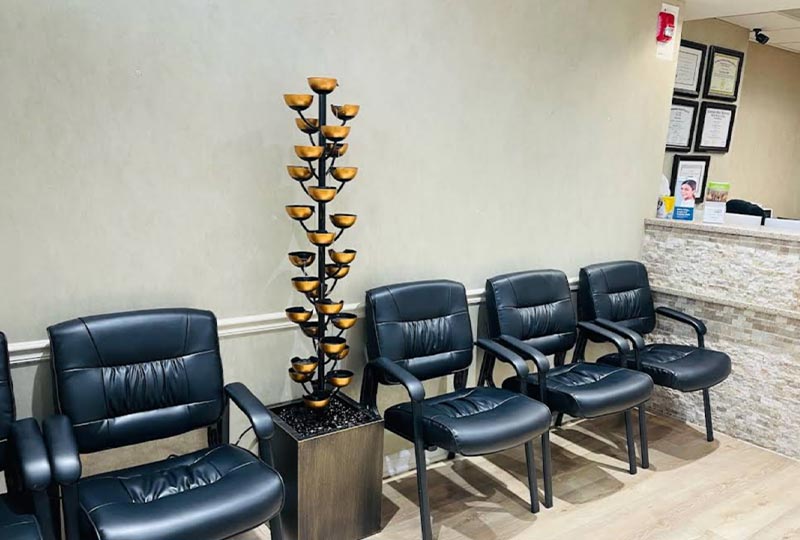 The image shows an interior view of a waiting area with multiple chairs, a reception desk, and a tall decorative plant.