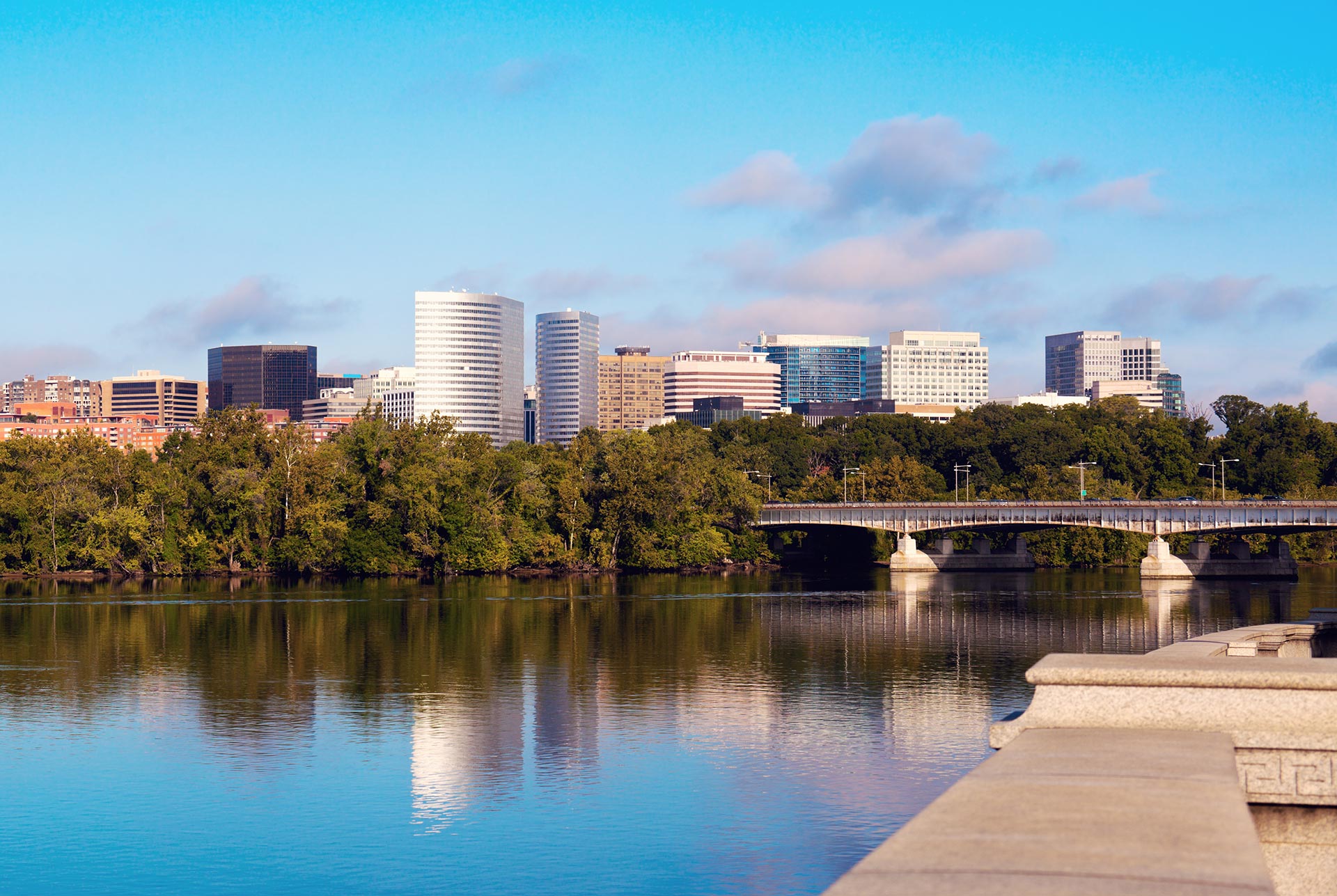 The image depicts a serene riverside scene with a city skyline featuring tall buildings, a bridge, and a clear blue sky reflecting off the water.