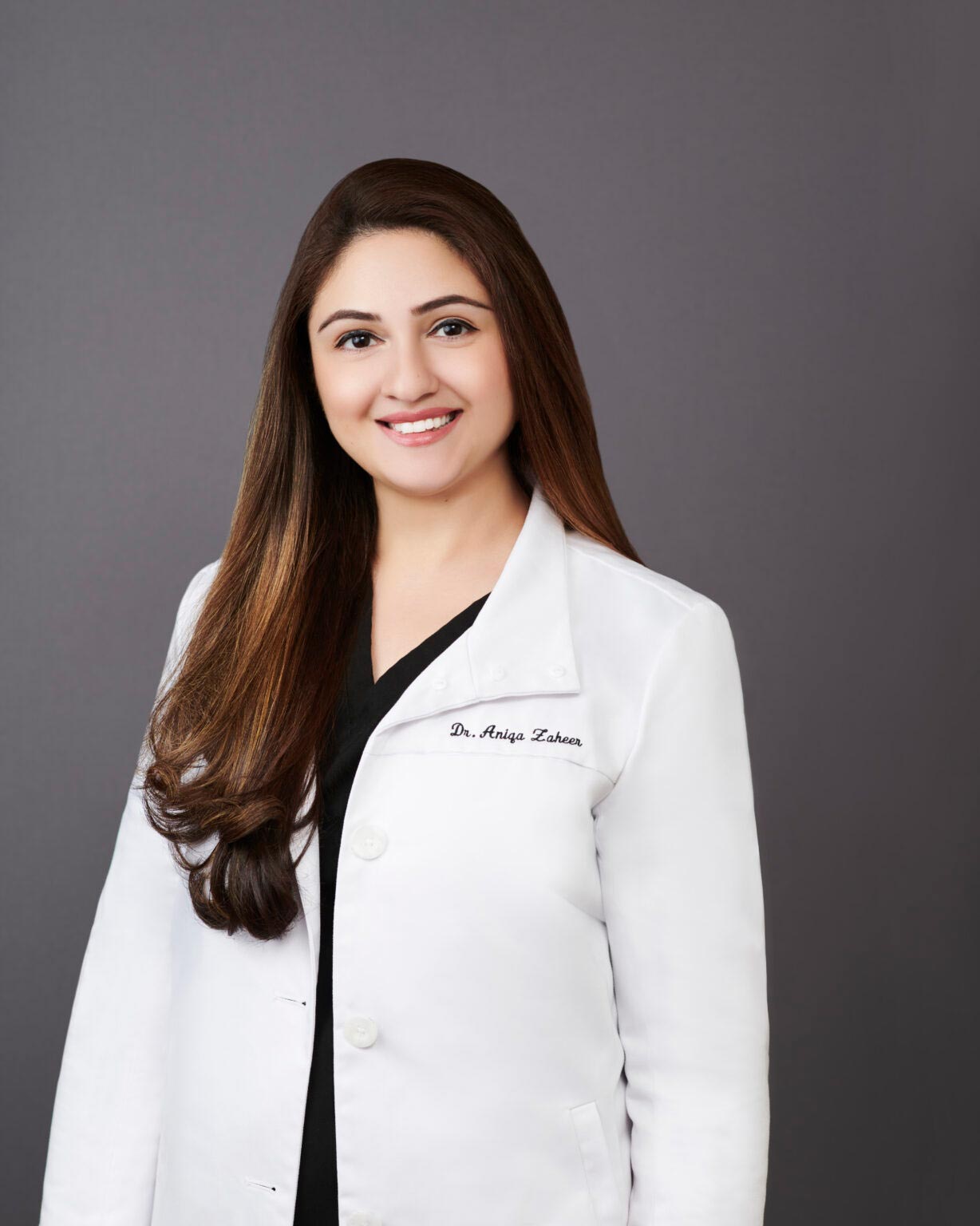 The image shows a woman wearing a white lab coat with a name tag on the left side of her chest, posing for a professional portrait against a neutral background.