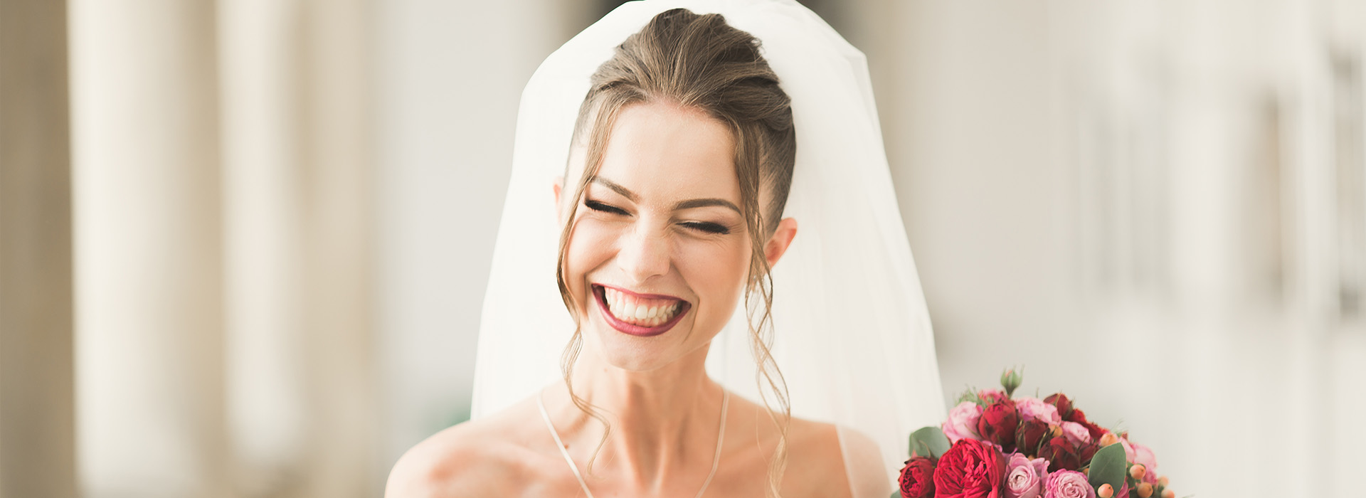 A bride wearing a white wedding dress and veil, smiling and holding flowers, with a groom behind her.
