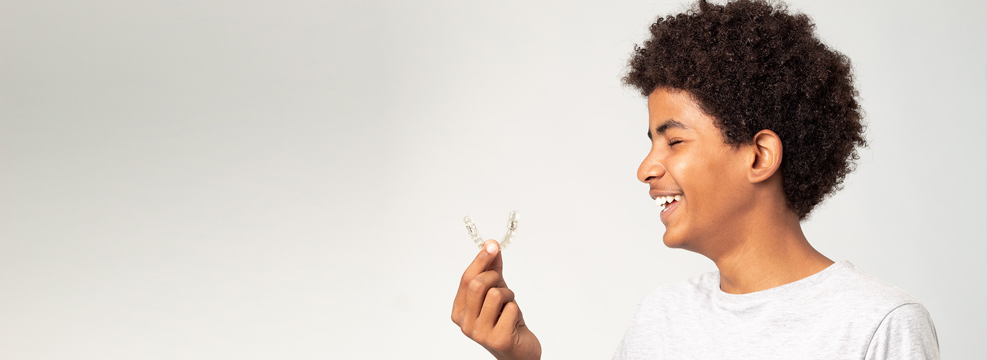 The image shows a young person with curly hair and a smile, holding a small object up to their face, set against a plain background.