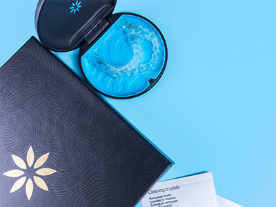 The image shows two closed cosmetic containers with intricate designs, placed on a surface against a blue background, accompanied by an open product with a visible logo, and a small amount of a substance resembling a greenish-blue gel inside.