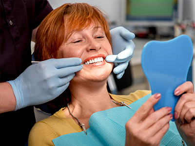 The image shows a woman sitting in a dental chair with a smile on her face, holding up a blue toothbrush with one hand while looking at it, with a dental professional standing behind her, examining her teeth with a mirror and wearing gloves.