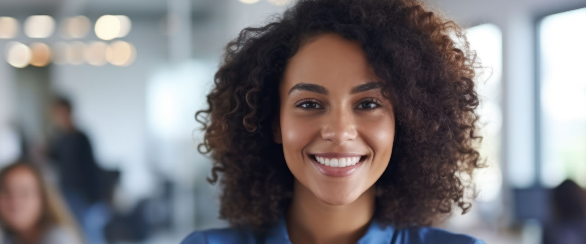 A smiling woman with curly hair, wearing a blue shirt, stands confidently in front of an office setting.