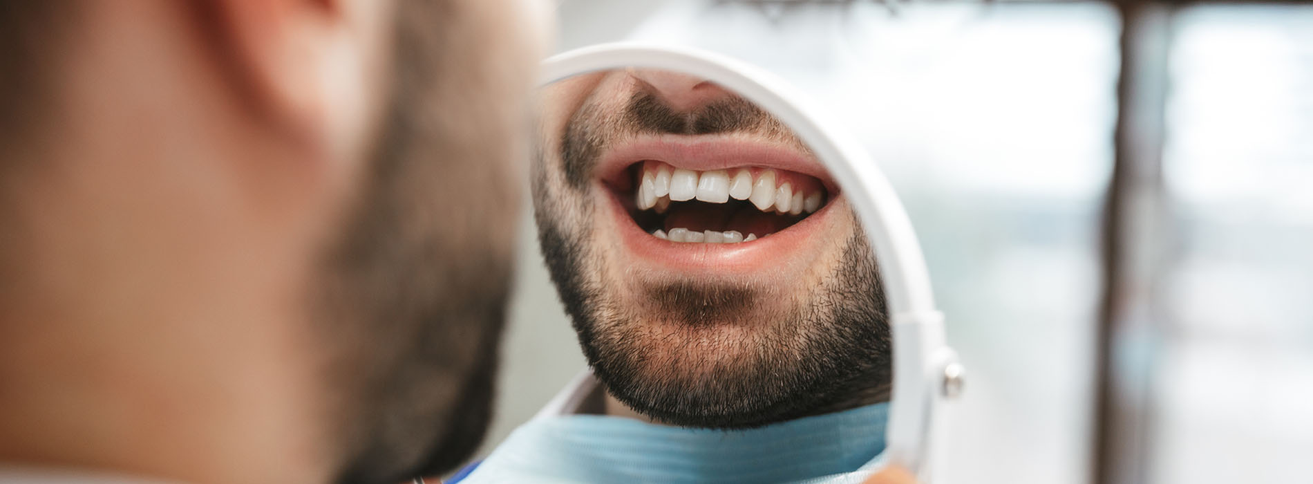 The image shows a close-up of a man with facial hair smiling at the camera, his mouth open slightly, and he appears to be wearing a surgical mask.