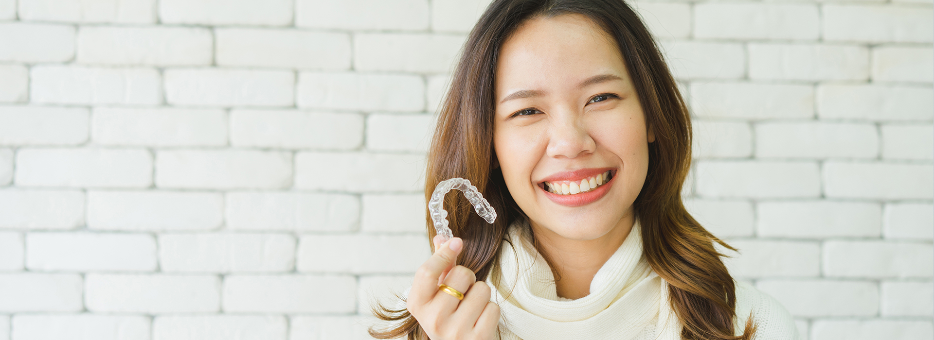 A woman with a smile holding up a toothbrush against her teeth, standing in front of a brick wall.