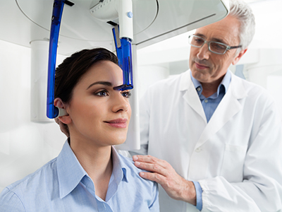 A woman wearing a blue headset while a man in a lab coat stands behind her, both are in a medical setting with an MRI machine.