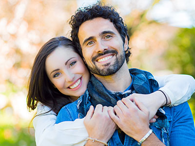 A man and woman embracing each other outdoors with trees in the background.