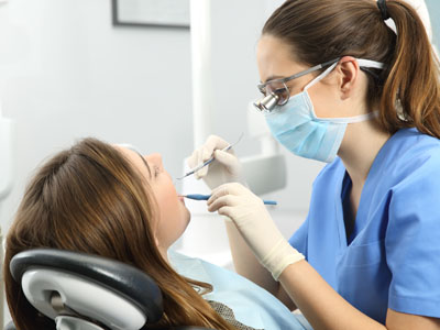 Woman wearing blue surgical mask performing dental procedure on patient s teeth, with dental chair and tools visible.