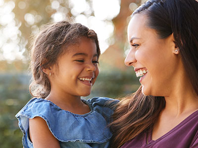 A woman and a young child are smiling at each other outdoors during the daytime.