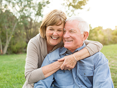 An elderly couple, a man and woman, are hugging each other outdoors during daytime.