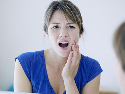A woman with her mouth open, holding her hand to her face, looking into a mirror while expressing concern or surprise.