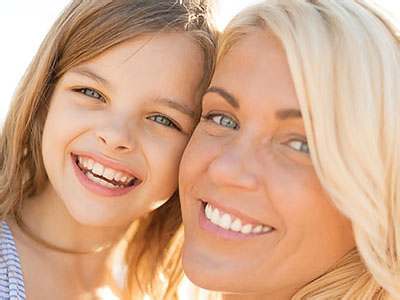 The image shows a woman and a young girl smiling together  they appear to be enjoying a sunny day outdoors, with the woman s face partially obscured by sunglasses.