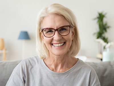 The image shows a woman with short blonde hair wearing glasses and a grey top, smiling at the camera.
