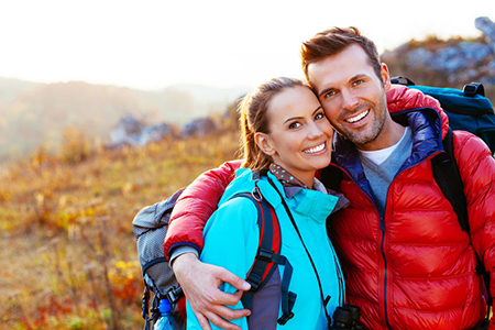 A man and woman smiling together in an outdoor setting, posing with backpacks and hiking gear, suggesting a shared adventure or exploration.