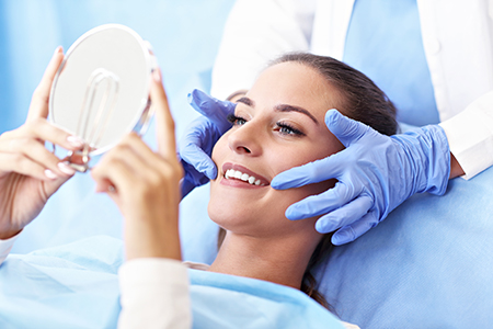 A woman sitting on a dentist s chair with a mirror held up to her face, smiling and looking at her reflection, while a dental professional adjusts a device on her head.