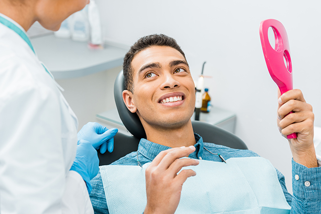 The image shows a person sitting in a dental chair with a smiling expression while holding a pink object, possibly an oral hygiene device, with a dental professional standing nearby wearing a white coat and face mask.