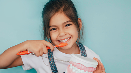The image shows a young girl brushing her teeth with a large toothbrush while smiling at the camera.