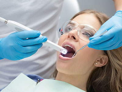This is a color photograph showing a woman sitting in a dental chair with her mouth open, receiving dental care from a professional using a device that appears to be for oral examination or cleaning. The setting suggests a dental office environment, and the woman is wearing blue gloves, which are commonly used by dental professionals during procedures.