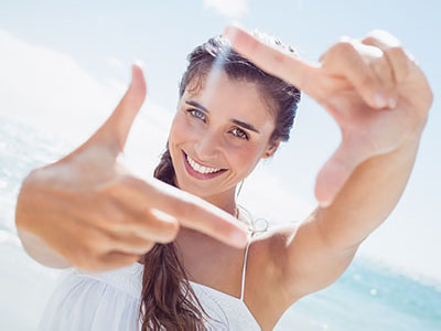 The image shows a young woman with long hair smiling at the camera while holding up a peace sign with both hands. She is outdoors, wearing a white top, and appears to be enjoying herself on a sunny day.
