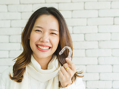 A young woman with dark hair is smiling at the camera while holding up a toothbrush. She stands against a brick wall backdrop, and there s another person partially visible on the right side of the image.